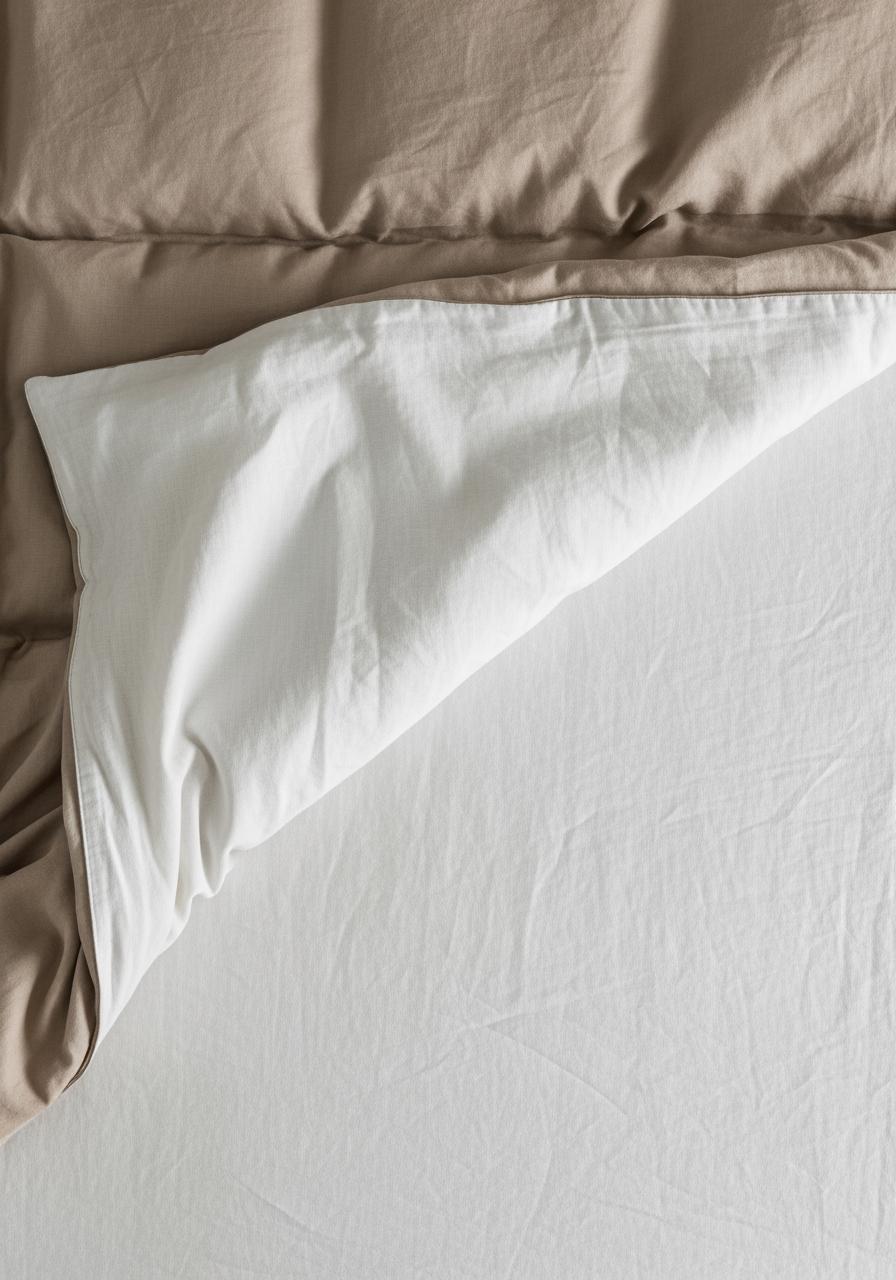 Vue de dessus d'un lit avec la housse de couette retirée en diagonale pour révéler un drap en lin texturé, illustrant les couches de linge de lit.